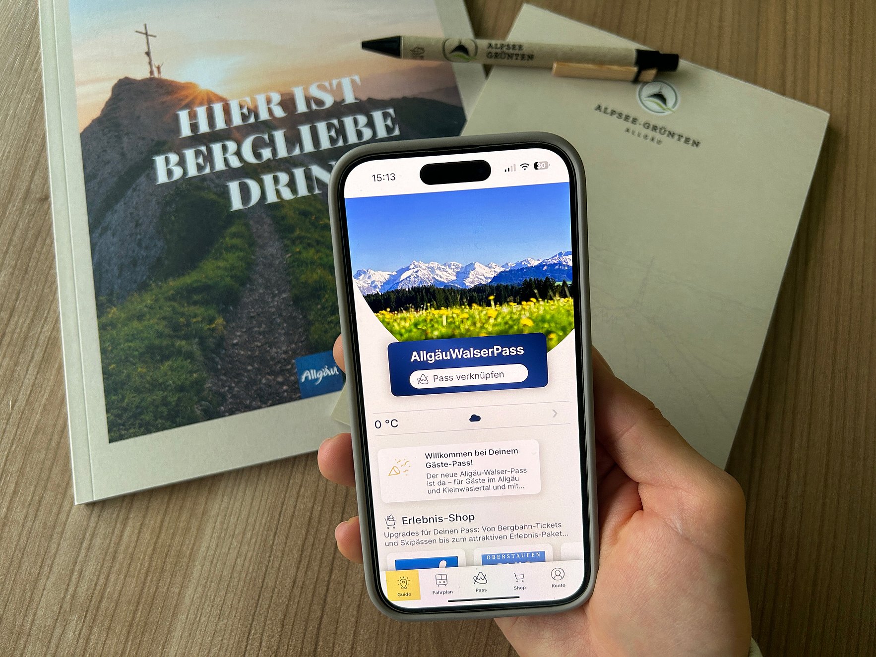Das Bild zeigt eine menschliche Hand mit einem Smartphone. Auf dem Smartphone ist der digitale Gästepass "Allgäu-Walser-Pass" zu sehen. Im Hintergrund liegt Werbematerial auf dem Schreibtisch.